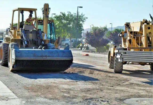 Two construction vehicles work on a road under repair, with a worker operating one machine and loose dirt visible on the ground.