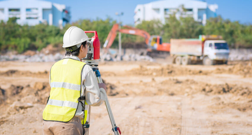 A construction worker in a safety vest and helmet uses surveying equipment at a construction site with machinery and trucks in the background.
