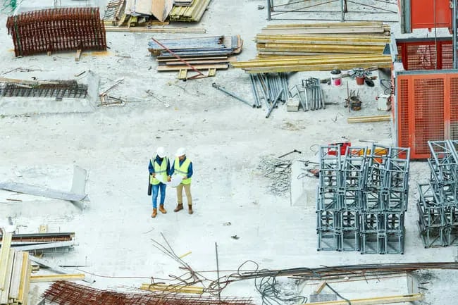 Two construction workers wearing safety vests and helmets stand on a building site surrounded by stacks of metal, wood, and construction materials.
