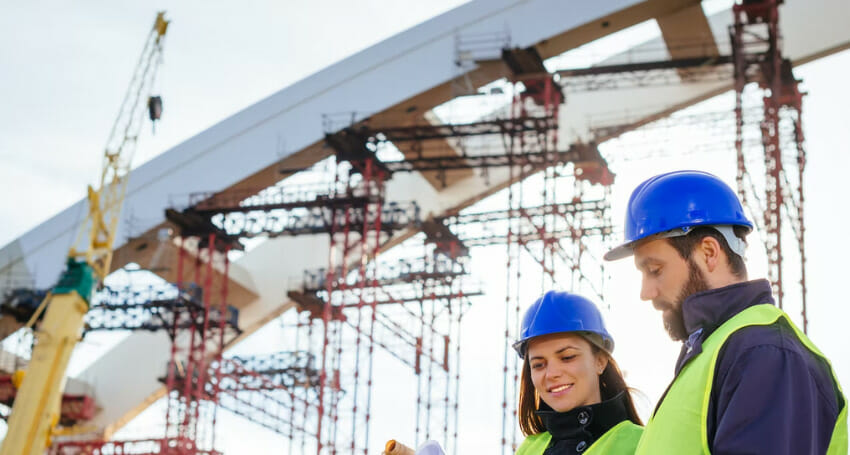 Two construction workers in safety vests and blue helmets review documents at a building site with scaffolding and a large arch structure in the background.