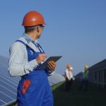 Young Korean man in overall and hardhat using tablet in sunlight standing on field with solar panels