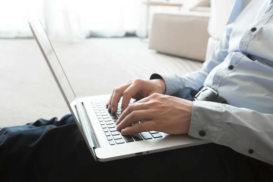 Close-up of a person in a light blue shirt typing on a laptop while sitting on the floor indoors, utilizing Copilot for Microsoft 365.