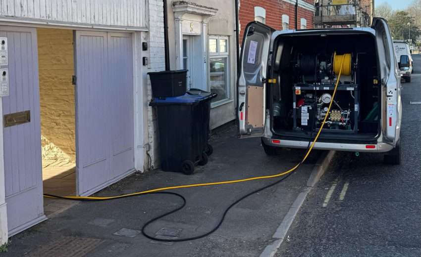 A drainage engineer in Portsmouth performing high-pressure water jetting on a residential driveway during winter with visible hose, safety gear, and water spray.
