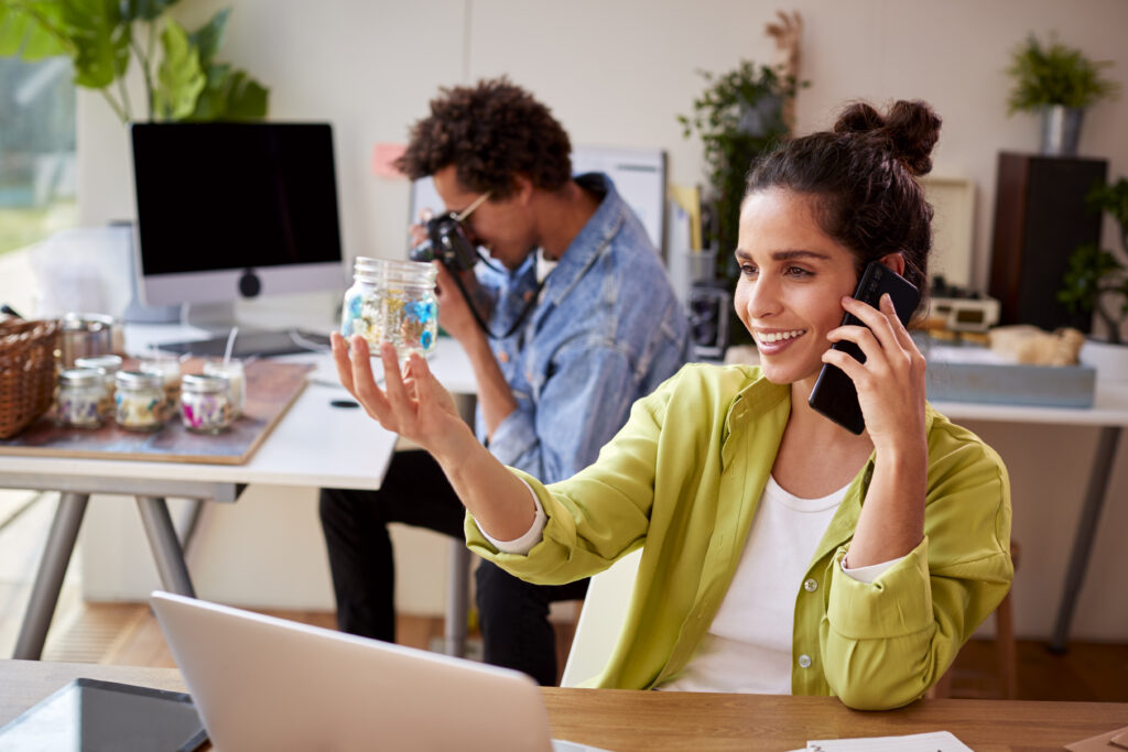 A woman in a bright green shirt smiles while talking on a smartphone and holding a jar of colorful paper stars, with a male colleague working at a computer in the background in a modern office setting.
