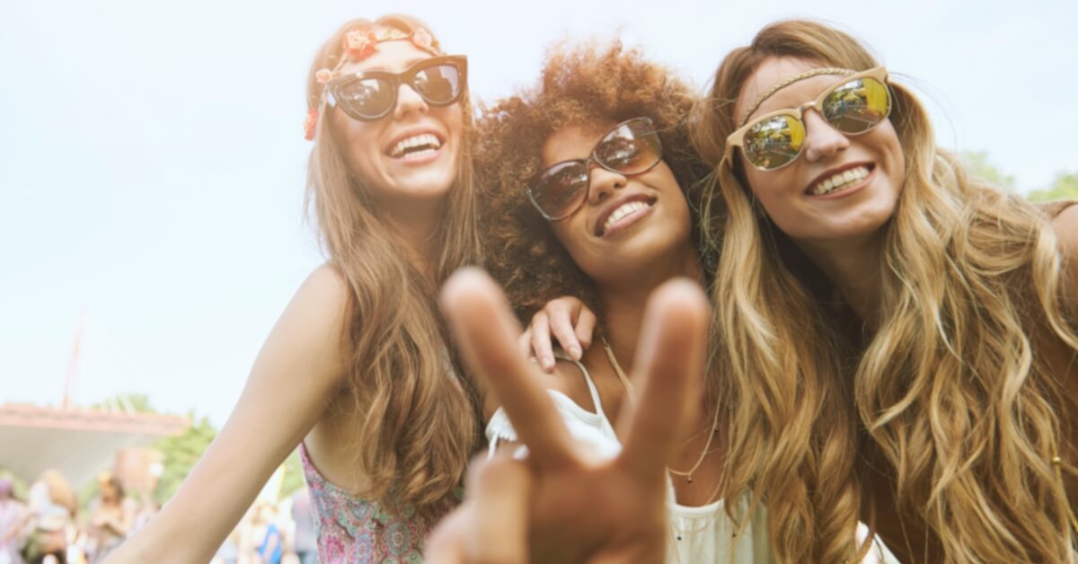 Od chronicznego stresu do dobrostanu. Trzy osoby w okularach przeciwsłonecznych i swobodnych strojach uśmiechają się, robiąc sobie selfie na zewnątrz. Jedna osoba wykonuje gest znaku pokoju.