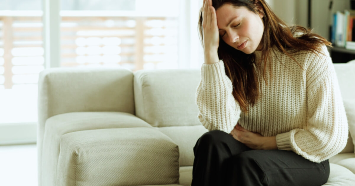 Od chronicznego stresu do dobrostanu. Kobieta siedzi na beżowej sofie, trzymając się jedną ręką za głowę, a drugą za brzuch. Wygląda na chorą.