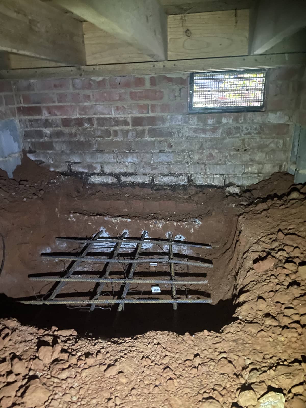 A dirt hole with a metal rebar grid at the bottom is dug under a brick wall in a crawl space, with a vent in the wall above.