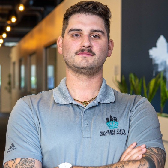 A man with short hair and a mustache stands with arms crossed, wearing a gray Queen City Construction and Design polo shirt in a modern office hallway.