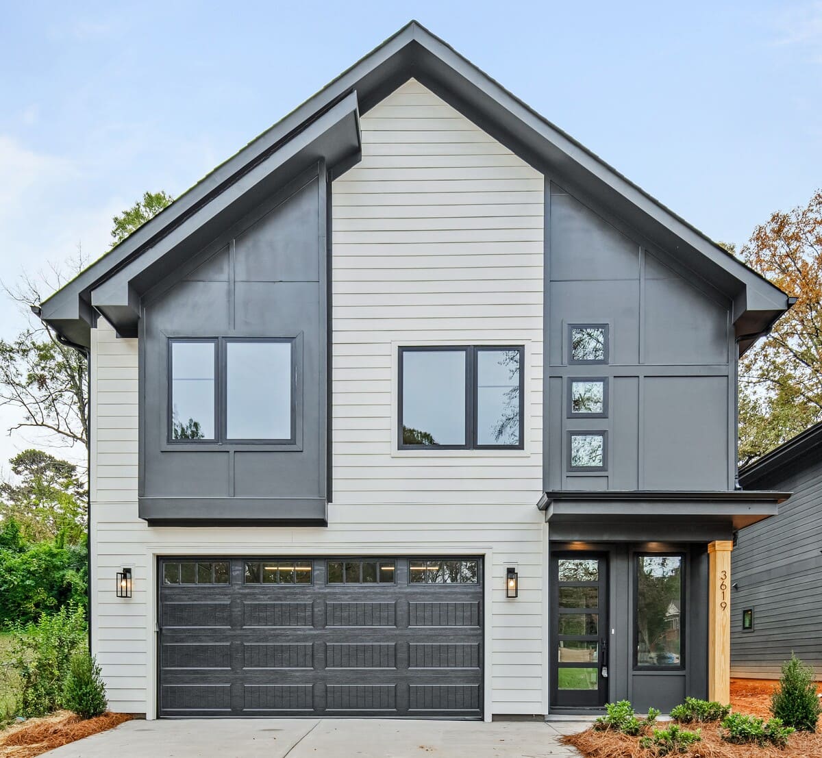 Modern two-story house with gray siding, black-framed windows, and a double garage door. The entrance is on the right side with a paved driveway and minimal landscaping.