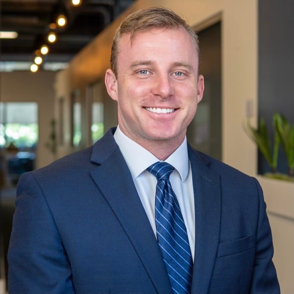 A man in a blue suit and striped tie smiles while standing in a modern office hallway with plants and windows in the background.