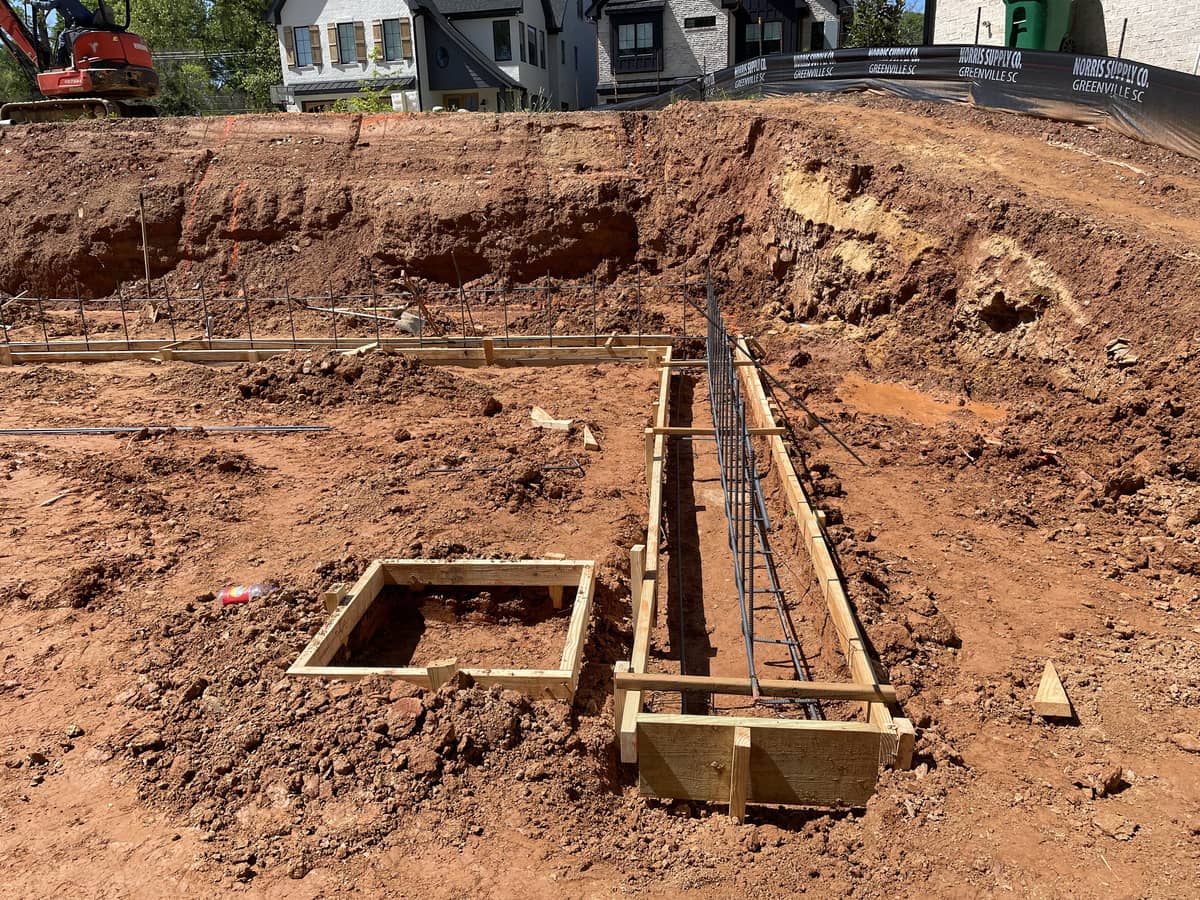 Construction site with exposed red dirt, wooden forms, and steel rebar set for concrete foundation work; houses and construction equipment visible in the background.