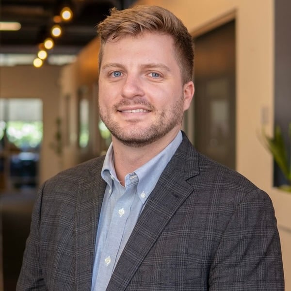 A man with short blond hair and a beard, wearing a gray blazer and light blue shirt, stands indoors in a modern office hallway.