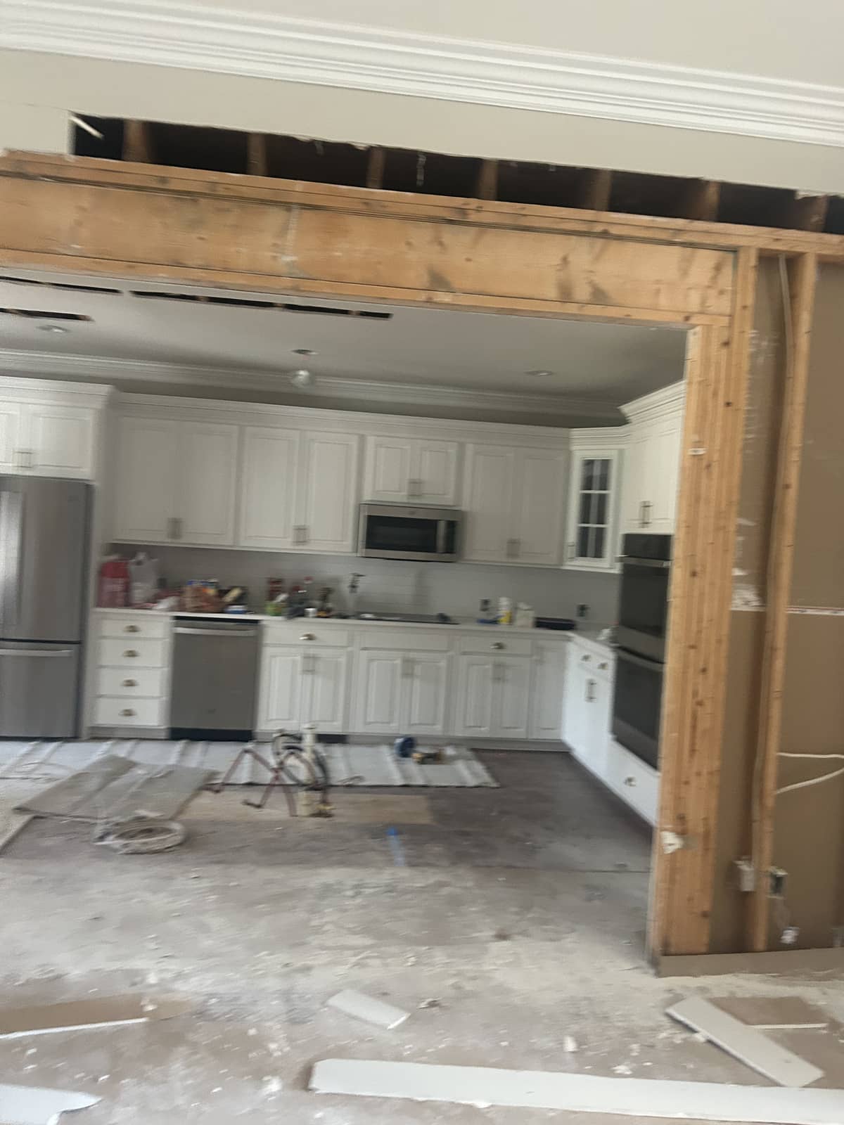 A kitchen with white cabinets and appliances is partially revealed through a large, unfinished wooden frame during home renovation. Construction debris is scattered on the floor.