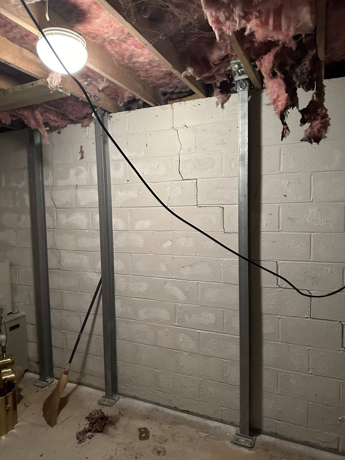 A basement wall with visible cracks is reinforced by three steel beams, with exposed insulation overhead and a light hanging from the ceiling. A broom leans against the wall.