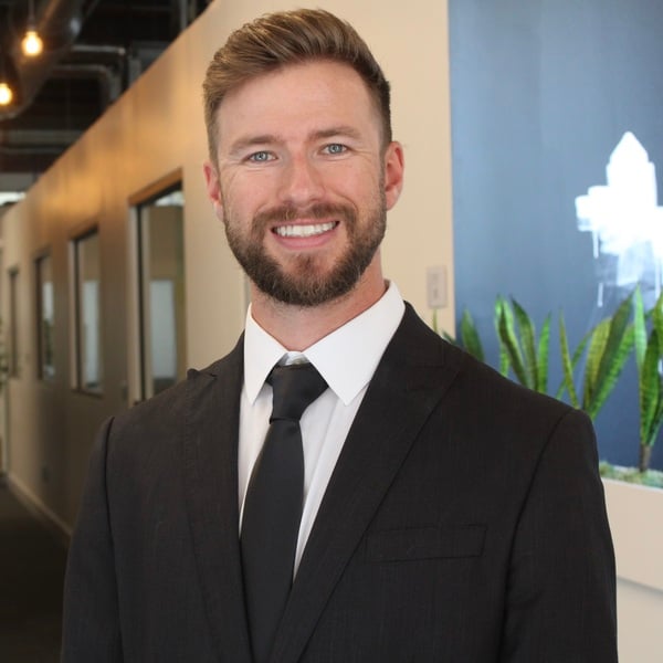 A man in a dark suit and tie stands in a modern office hallway, smiling at the camera. There are plants and a blue wall in the background.