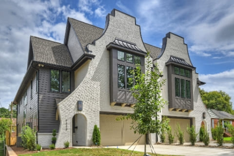 Two-story house with white brick facade, dark trim, large windows, steep gabled roofs, and a driveway, surrounded by greenery under a partly cloudy sky.