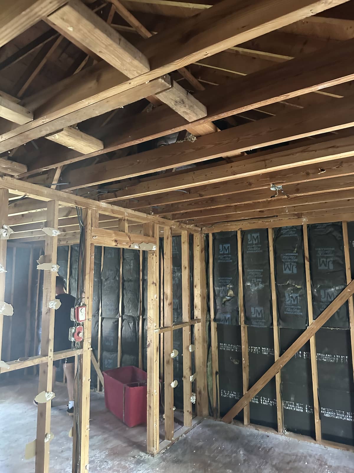 Interior view of a house under construction showing exposed wooden wall studs, ceiling joists, insulation, wiring, and a red storage bin on the floor.