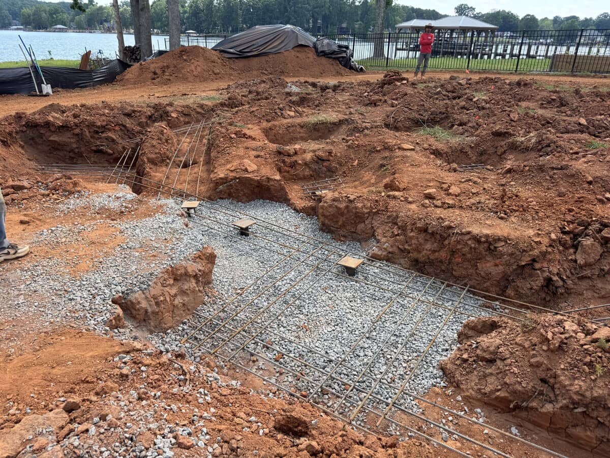 Construction site with exposed rebar and gravel in excavated trenches, piles of dirt nearby, and a lake with trees in the background.