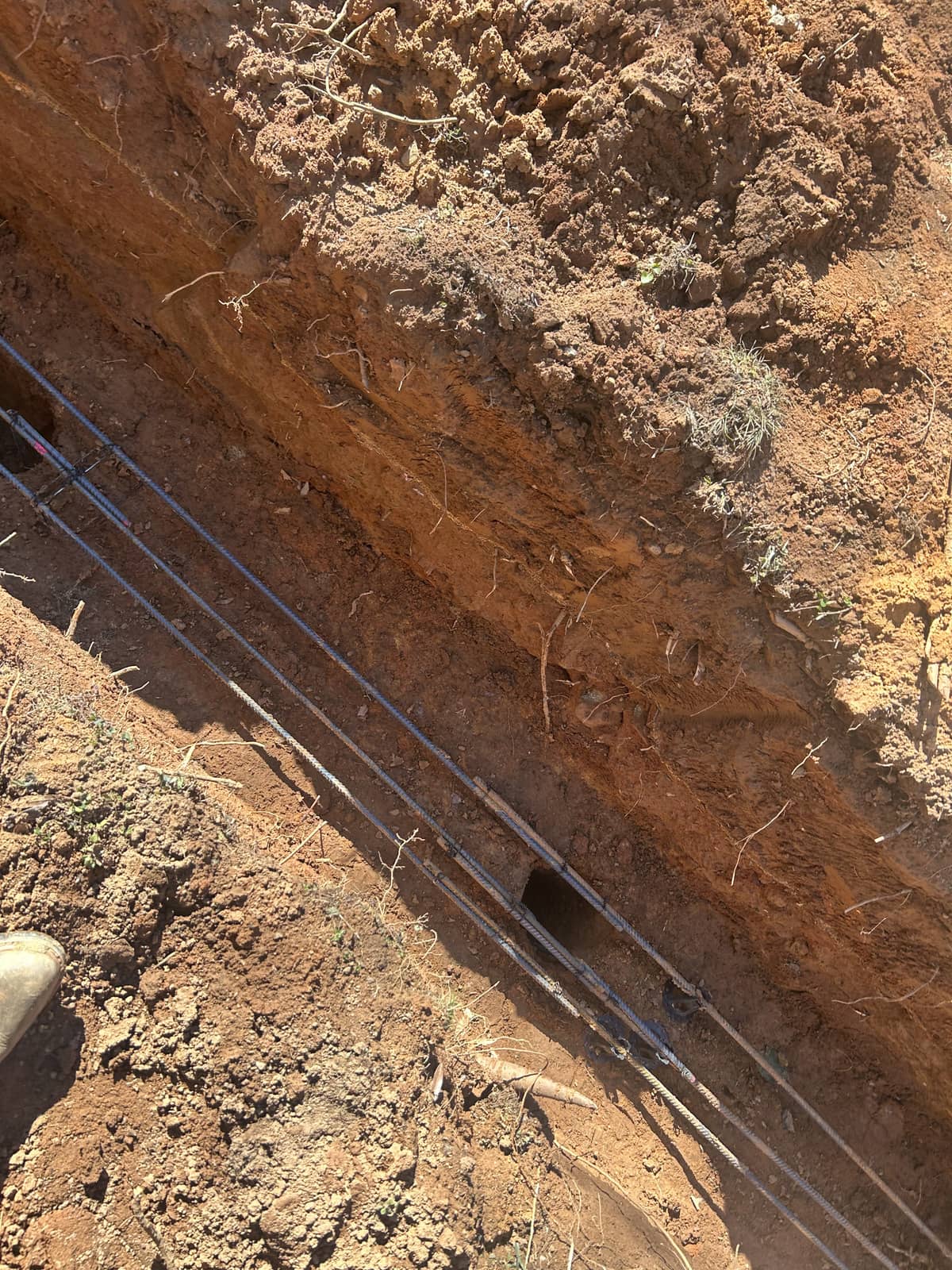 A trench dug in brown soil containing several metal pipes running parallel and a small dark hole in the trench wall.
