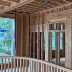 Wood framing of a house under construction with exposed studs, beams, and wiring visible; large windows face an outdoor view with trees.
