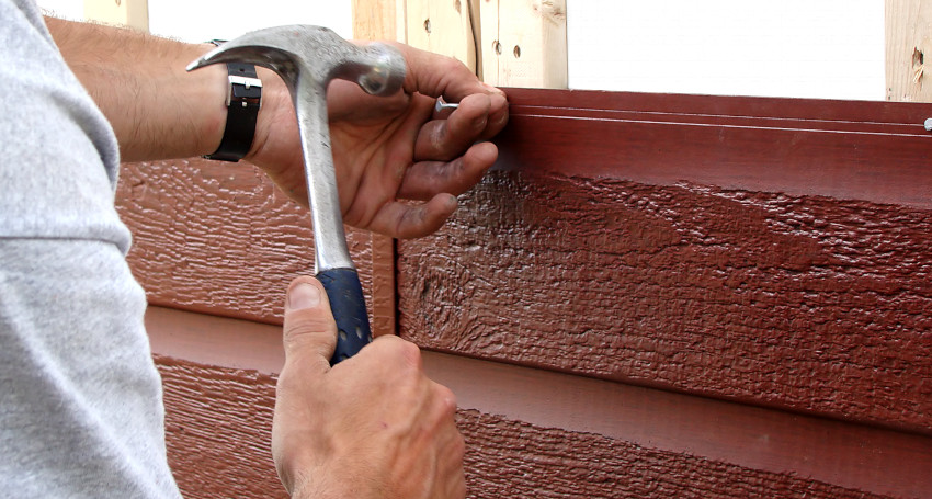 A person using a hammer to pull out a nail from the siding of a house.