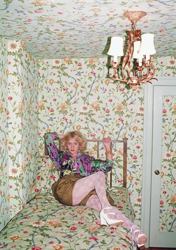 A woman sitting on a bed in a room with floral wallpaper, looking contemplative.