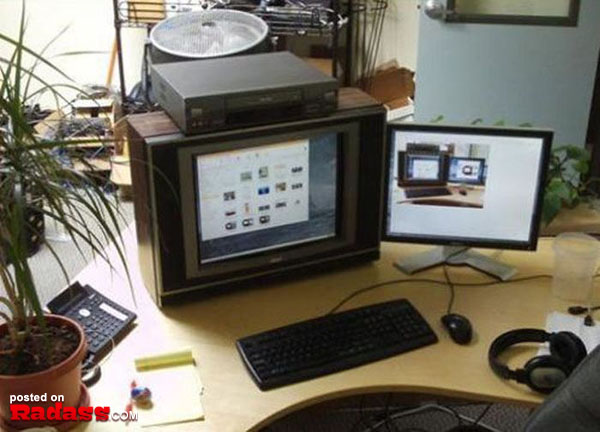 A desk with two monitors, a keyboard, and a plant adorned with 50 Redneck Remedies To Help You In Times Of Need book.