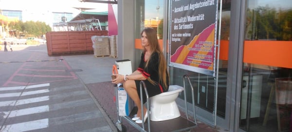 A woman is sitting on a cart in front of a store, but someone approaches her with an excuse -