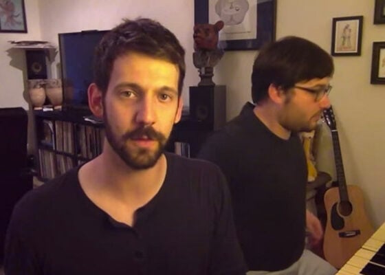 Two perfect men showcasing celebrity impressions next to a piano in a living room.