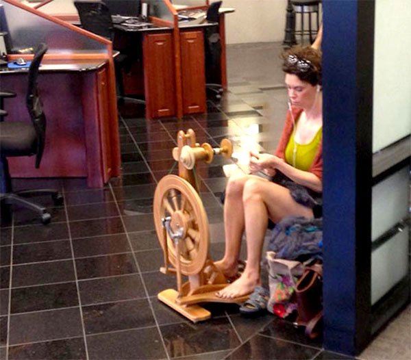 A woman sitting on a spinning wheel in an office. The office environment takes a surprising turn as a woman engages in the unconventional activity of spinning yarn while working. This unexpected blend of traditional craftsmanship