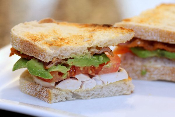 Food porn sandwich featuring chicken, tomatoes, and avocado on a pristine white plate.