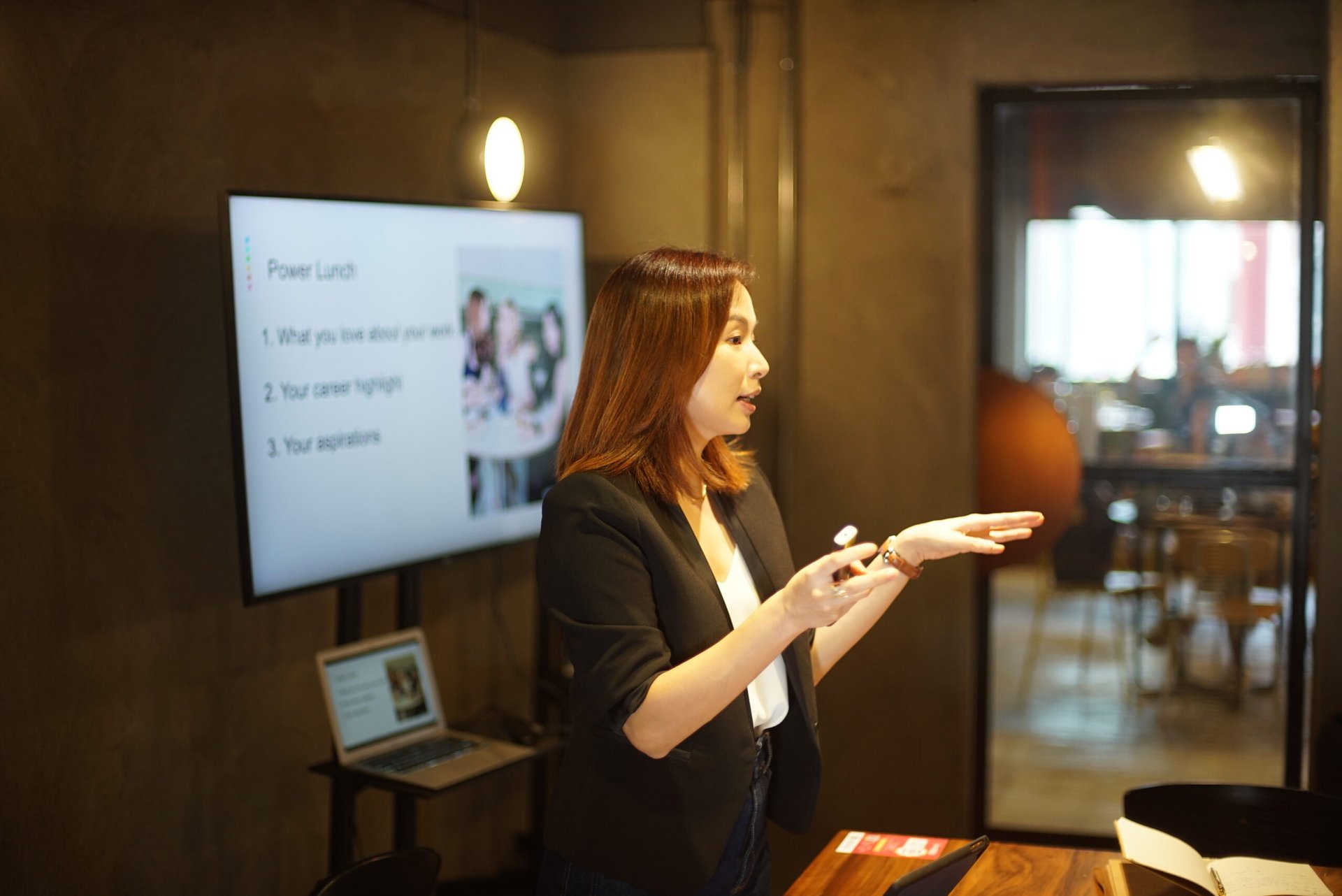 A person standing and speaking in a room, gesturing with one hand while holding a marker in the other. A screen beside them displays a presentation titled "Power Lunch" with three points listed, emphasizing peak performance and the benefits of a healthy diet.