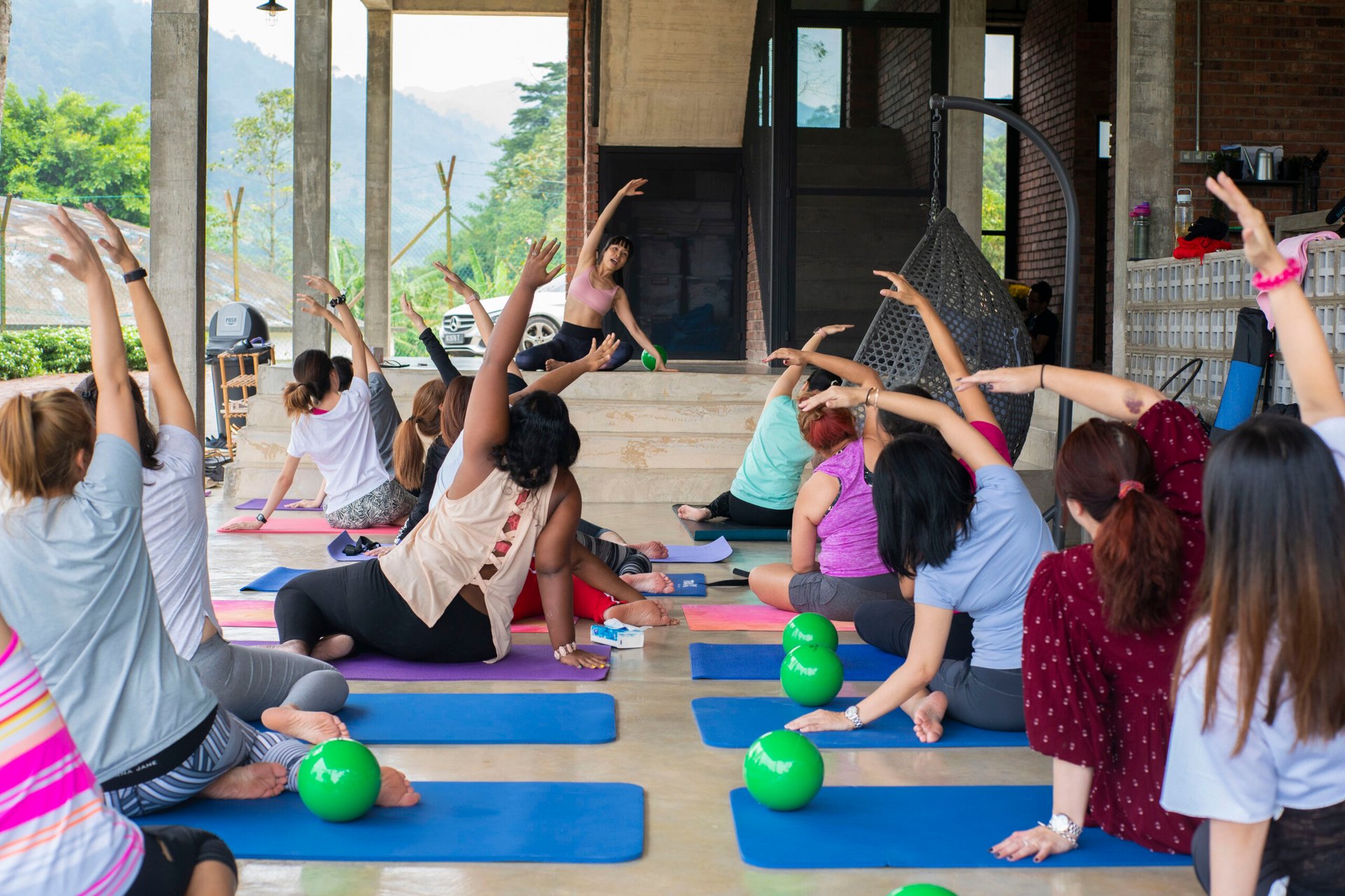 A group of people are participating in a yoga class outdoors, practicing stress management techniques while seated on colorful mats. Green exercise balls are placed nearby on the floor.