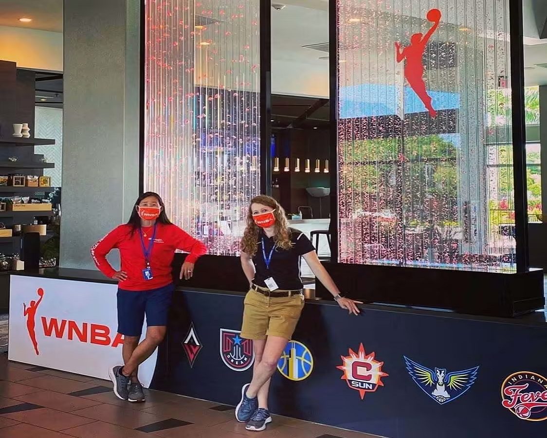 Women posing beside a vibrant indoor water feature with bubbles, featuring WNBA branding and logos of various basketball teams, in a modern mall setting.