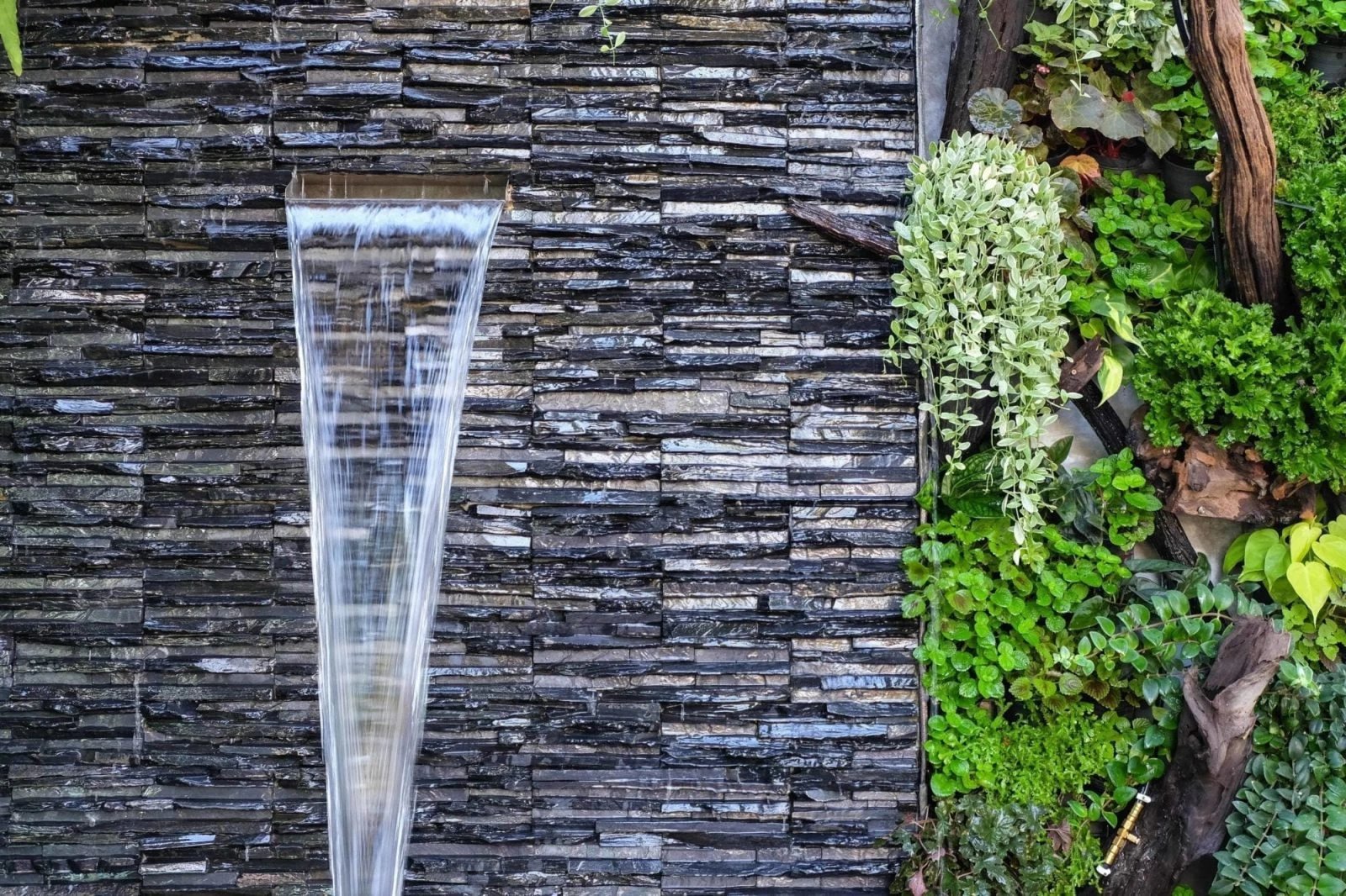 Indoor water wall feature with cascading water, surrounded by lush greenery and textured stone wall, enhancing ambiance in commercial spaces.