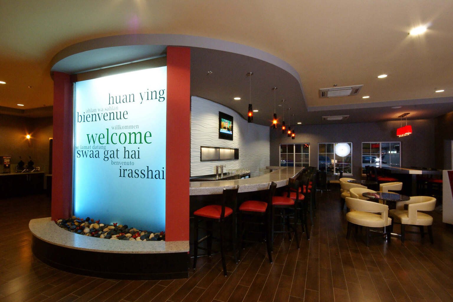 Interior of a modern hotel lobby featuring a welcoming wall with the word "welcome" in various languages, surrounded by decorative stones, sleek seating, and a bar area, emphasizing luxury and hospitality.