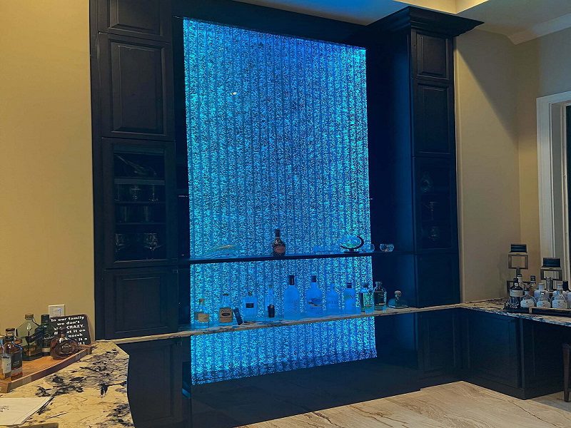 Indoor bubble wall water feature illuminated in blue, enhancing commercial space aesthetics, with a bar setup displaying various liquor bottles below.