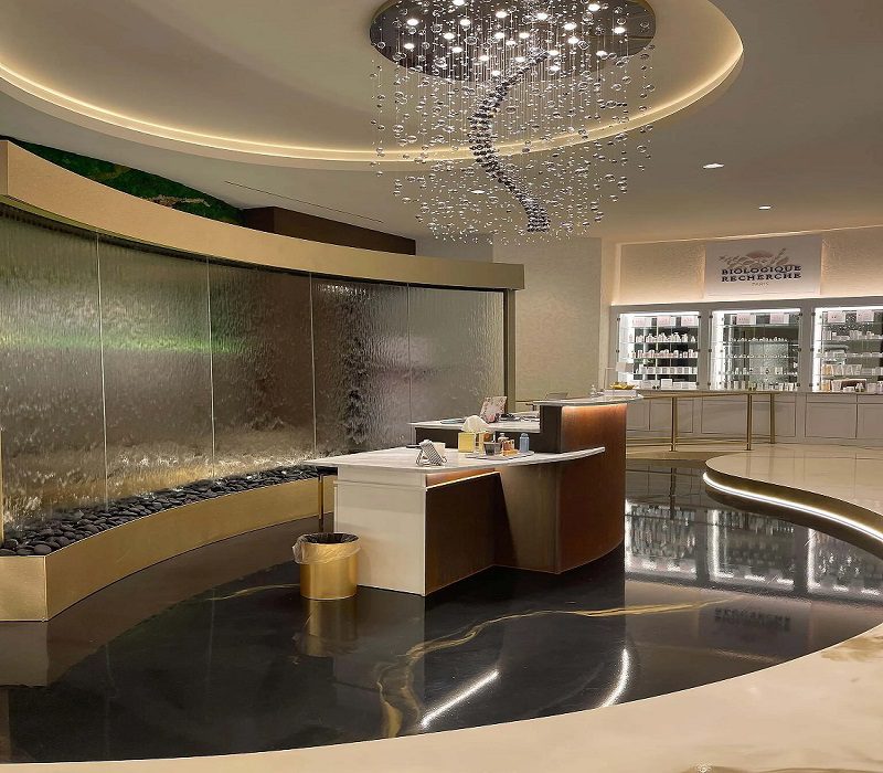 Indoor water feature with a curved glass wall, pebbles at the base, stylish reception desk, and modern lighting, enhancing the ambiance of a commercial space focused on wellness and design.
