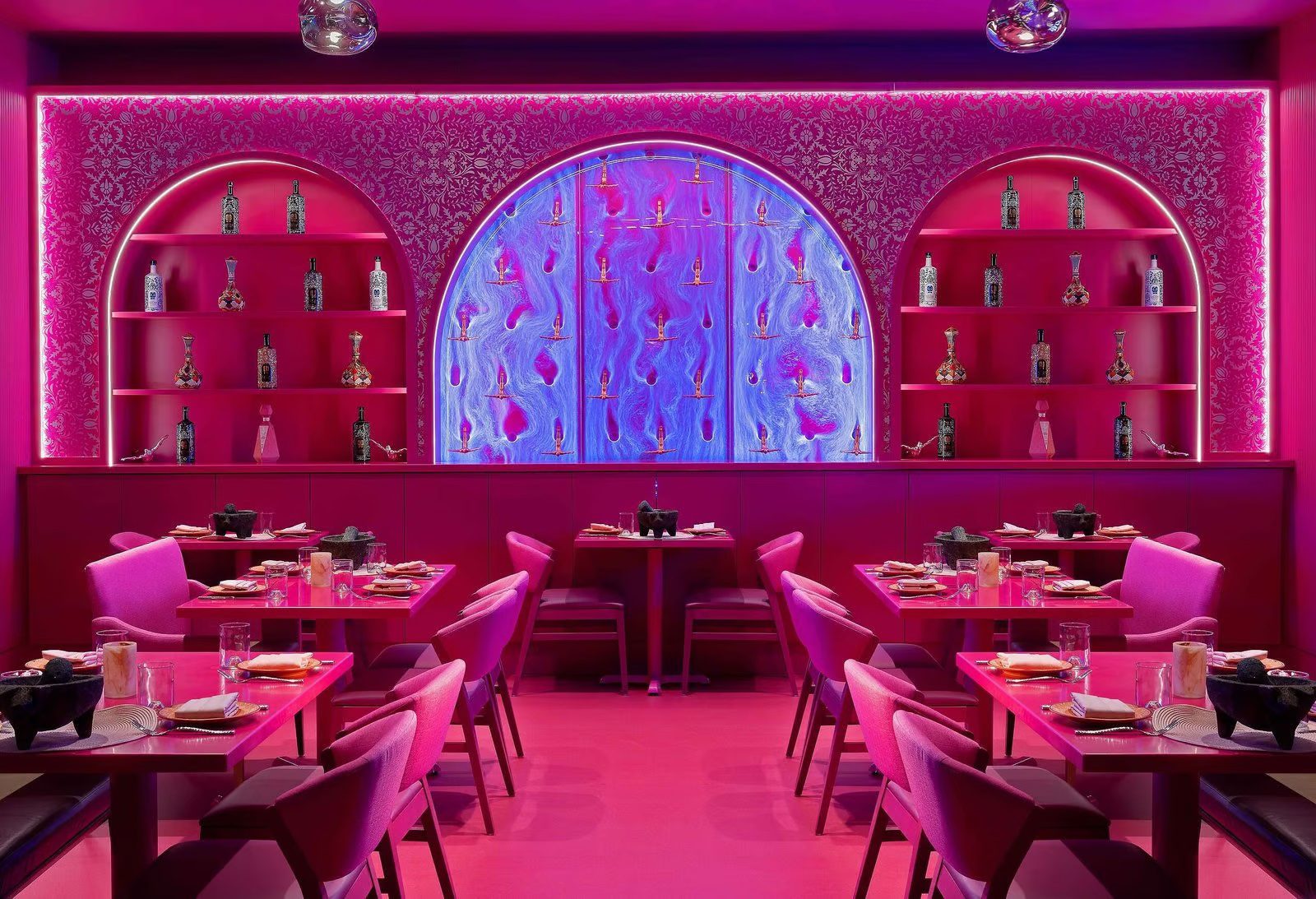 Indoor dining area featuring vibrant pink decor, illuminated water feature, and artistic display of bottles on shelves, enhancing the ambiance for restaurant patrons.