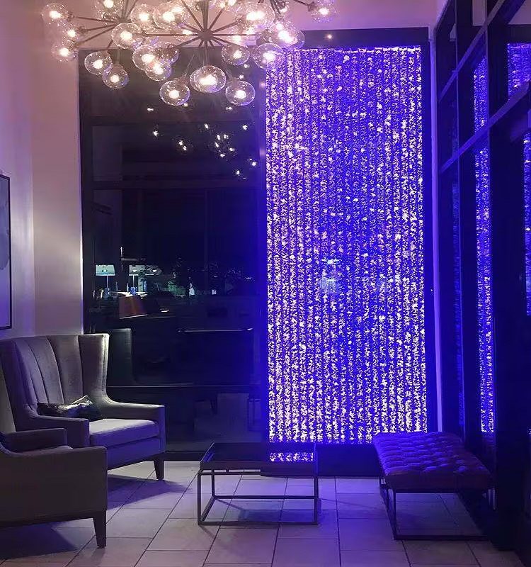 Indoor water bubble wall feature illuminated with purple lights beside modern seating area and chandelier, enhancing ambiance in commercial space.