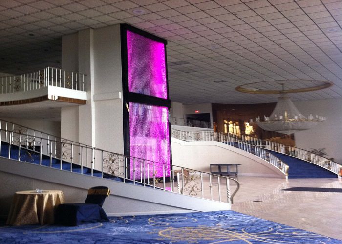 Indoor water feature with illuminated pink water curtain, enhancing the ambiance of a spacious commercial lobby with elegant staircase and chandelier.