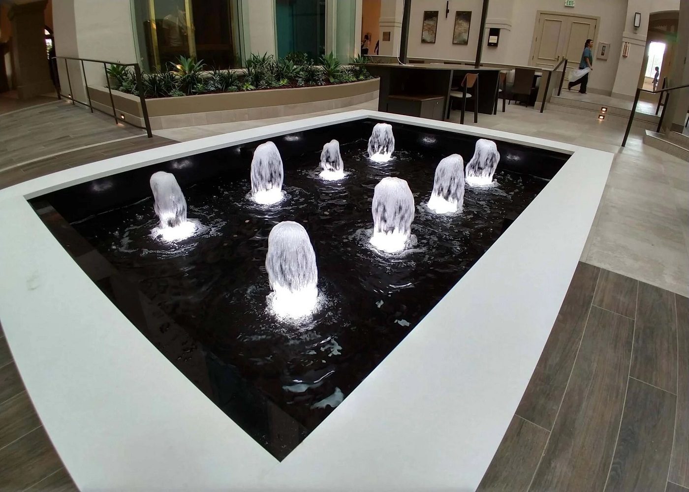 Illuminated water feature with multiple jets creating cascading water effects, surrounded by a sleek black basin and modern architectural elements, enhancing indoor commercial space aesthetics.