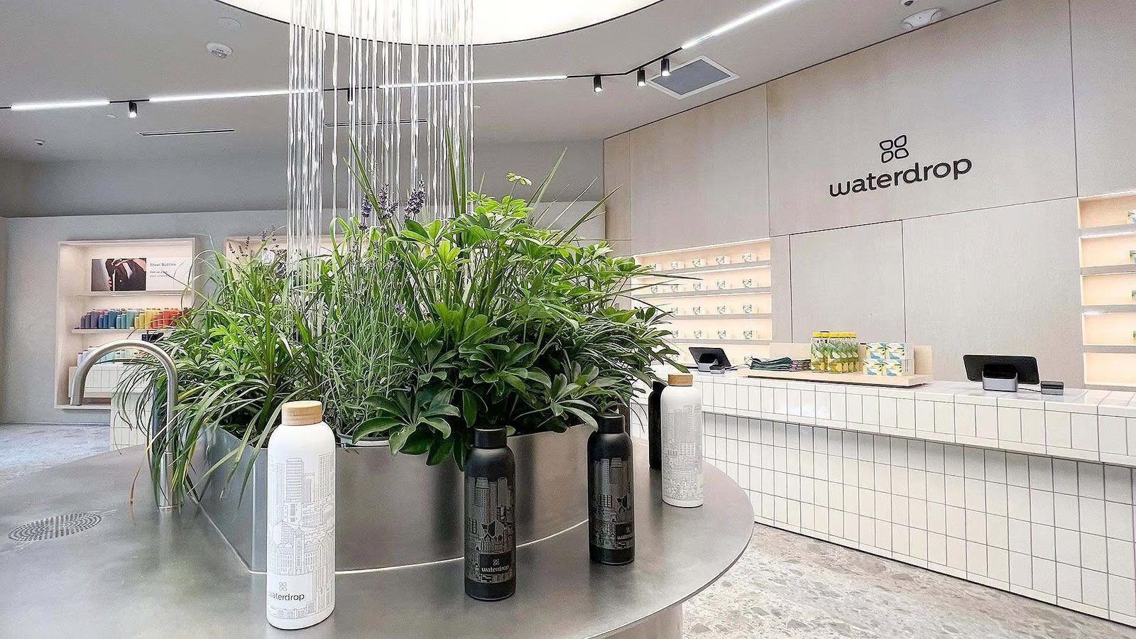 Indoor water feature with cascading water, lush greenery, and branded water bottles in a modern retail space.