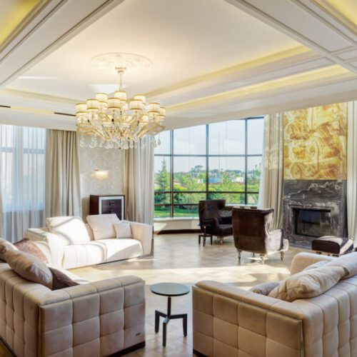 Luxury living room with big windows with panoramic view of garden and crystal chandelier in center of ceiling. Stylish interior of room with three sofas and armchairs around the fireplace. Photo of a high net worth living room - M&N Insurance