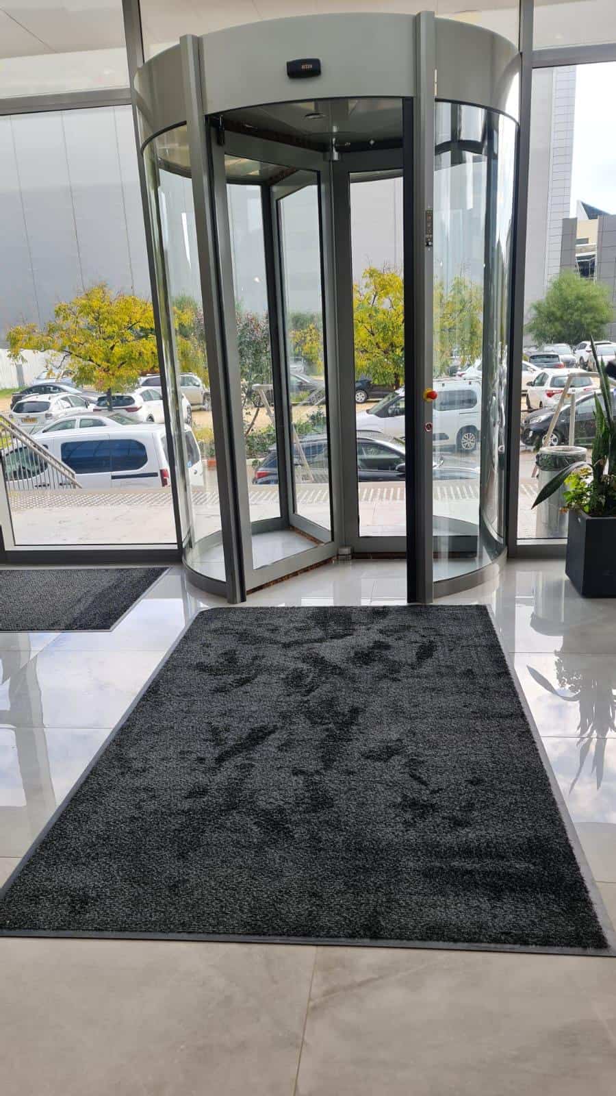 דלת זכוכית מסתובבת בכניסה לבניין כוללת שטיחי סף, עם מחצלת כהה על רצפה מבריקה ומכוניות ועצים הנראים מבחוץ.
