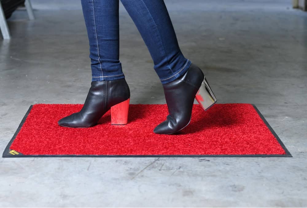אדם נועל מגפיים שחורים וג'ינס דורך על שטיח סף אדום מלבני, המונח על רצפת בטון. מתאים במיוחד בתור שטיח כניסה לעסק להגנה נגד החלקה.