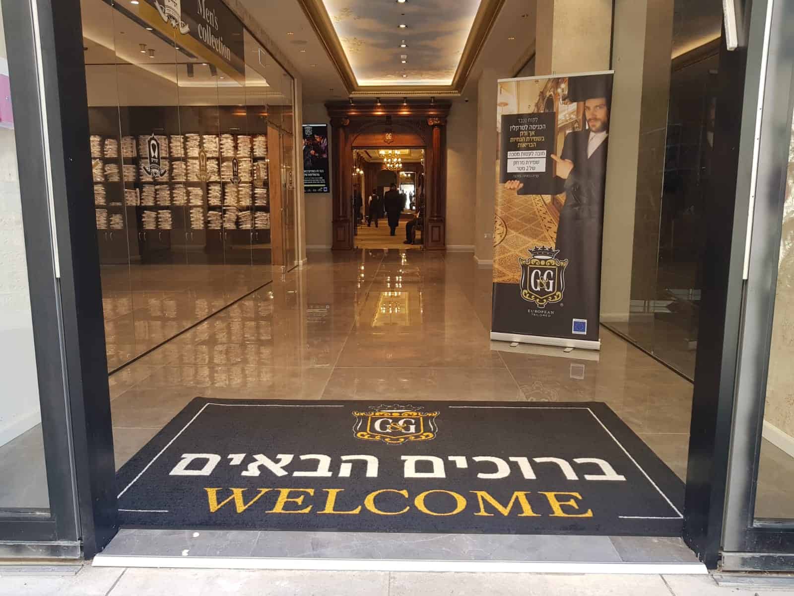כניסה לבניין עם שטיח שחור ועליו הכיתוב "ברוכים הבאים" באנגלית ובעברית; בפנים, מסדרון עם רצפת אריחים ושלט פרסומי מימין.