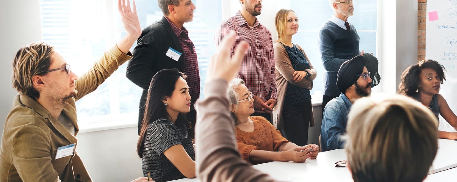 BIZ-LYNX Technology - A diverse group of people in a meeting room, with some participants seated and raising their hands, while others are standing and listening attentively. They appear to be engaged in a discussion about who we are as a team and our proven track record.