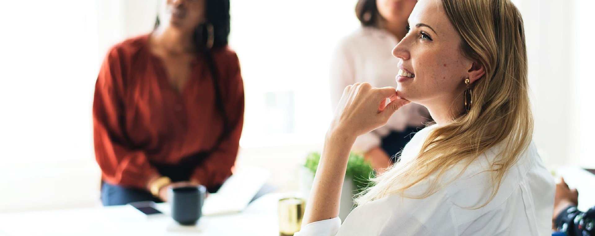 BIZ-LYNX Technology - A woman with blonde hair, dressed in white, sits attentively with her hand on her chin. In the background, two other people, slightly out of focus, sit and listen. Her posture reflects the essence of who we are—dedicated professionals committed to exceptional customer service.