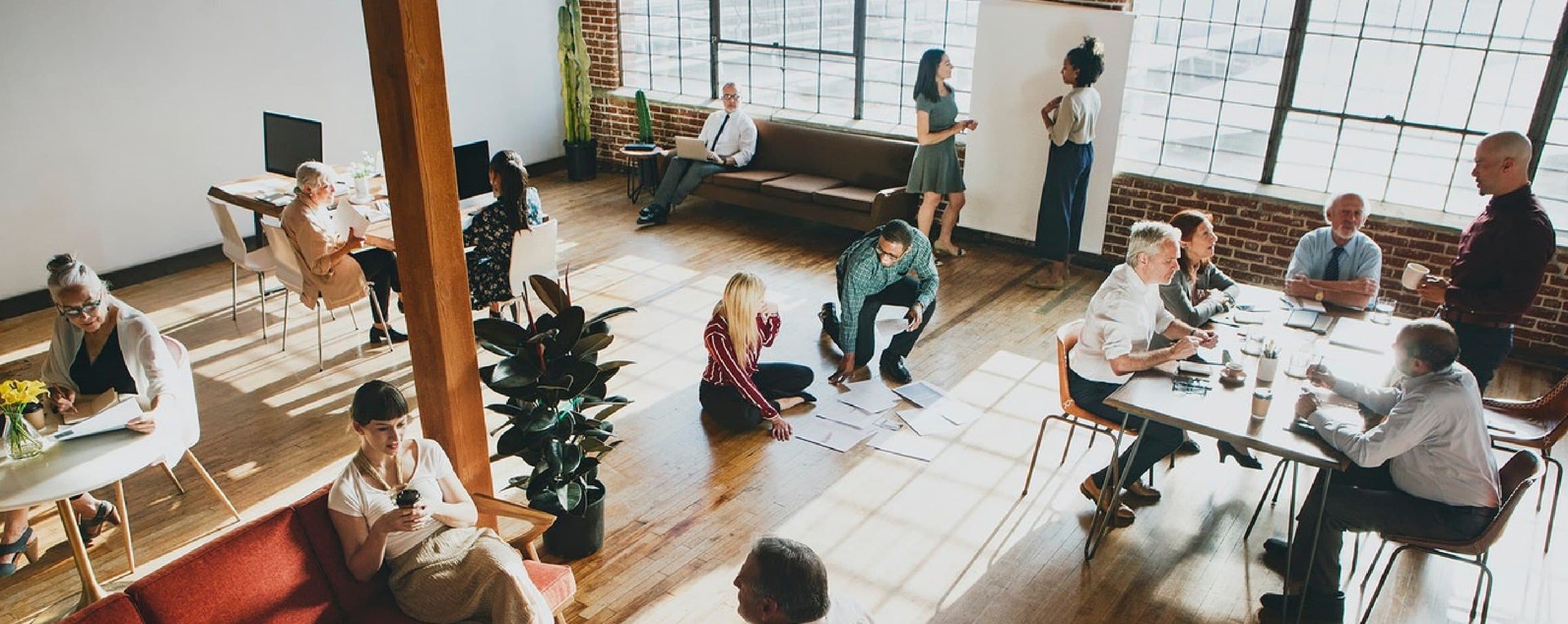 BIZ-LYNX Technology - A large room with people working and socializing, some sitting at tables, others standing or on the floor. Sunlight streams through large windows. The setting radiates a casual, collaborative atmosphere, reflecting who we are with our focus on exceptional customer service.